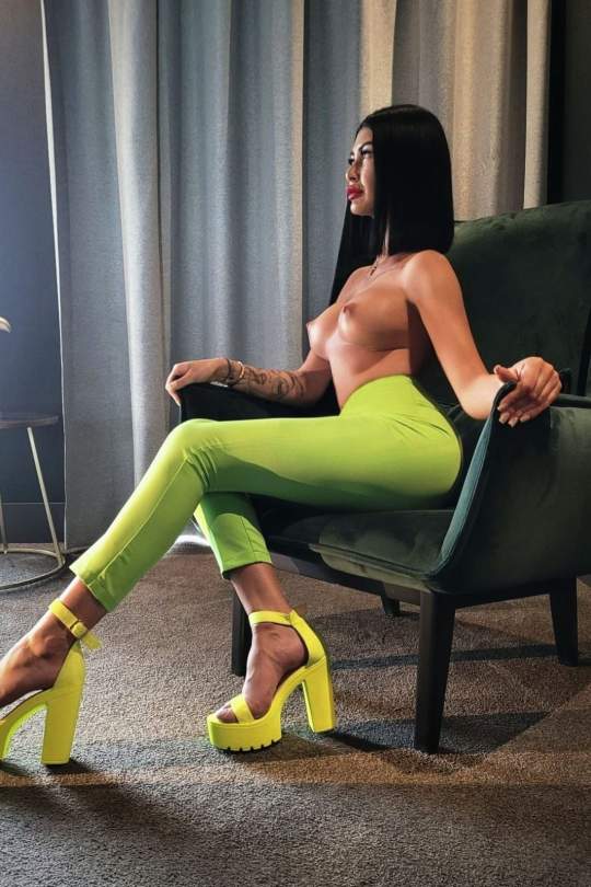 Patricia sitting while topless and wearing a pair of green trousers